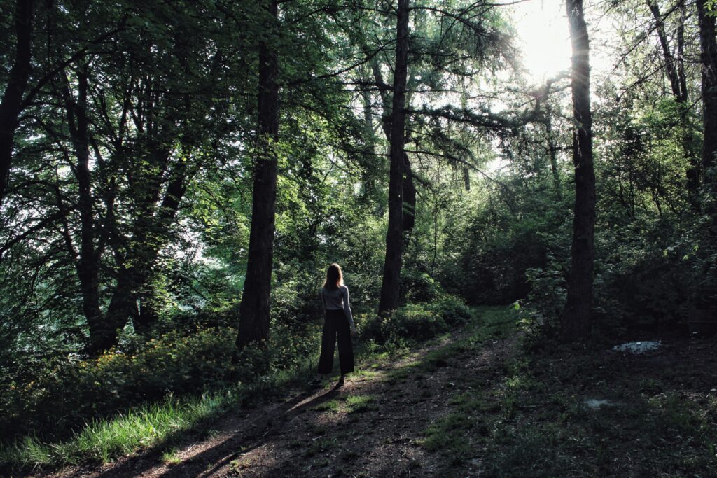 A woman walks alone through a sunlit forest pathway, surrounded by lush green trees.